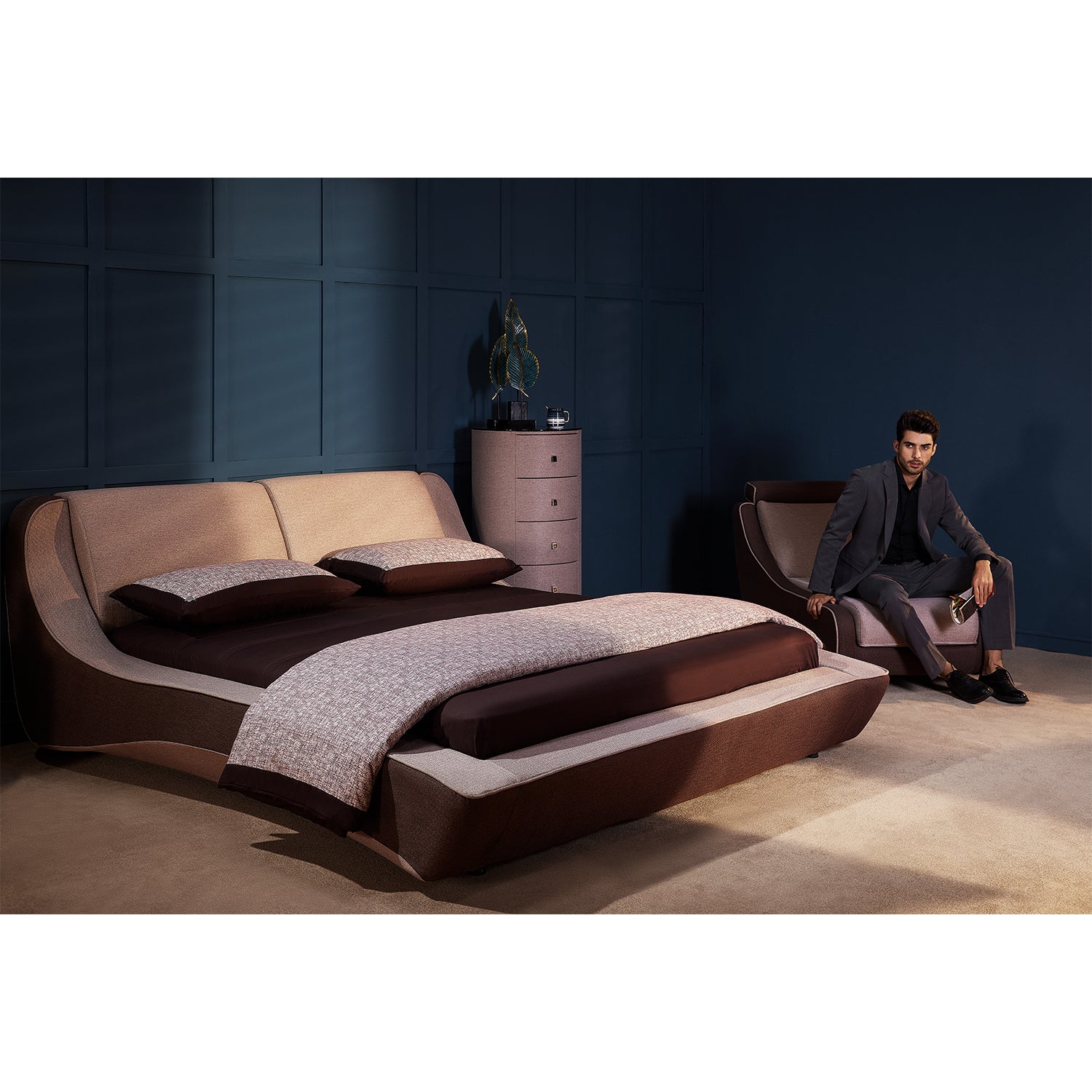 Modern bedroom with DeRUCCI brown bed frame inspired by sports cars, featuring contrasting colors and classic modern design. Matching chair and chest of drawers included in the background, with a man sitting in the chair.