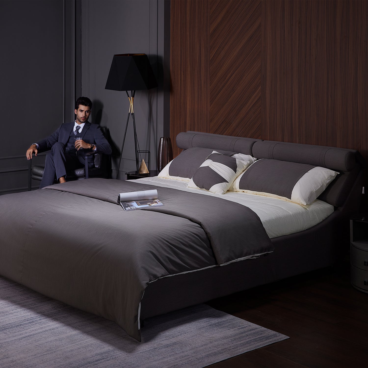 DeRUCCI Bed Frame BZZ4 - 093C with dark gray bedding and headboard, nightstand with lamp, and a man in a suit seated beside the bed in a modern bedroom setting.