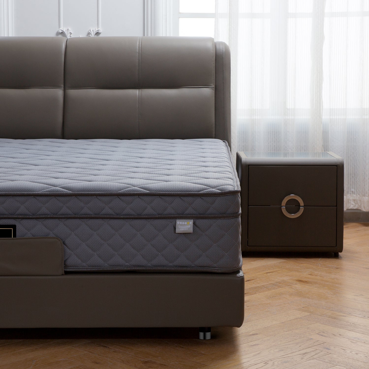 Minimalist bed frame with a padded leather headboard and grey quilted mattress, featuring a matching nightstand with circular handles, wooden floor, and sheer curtains.