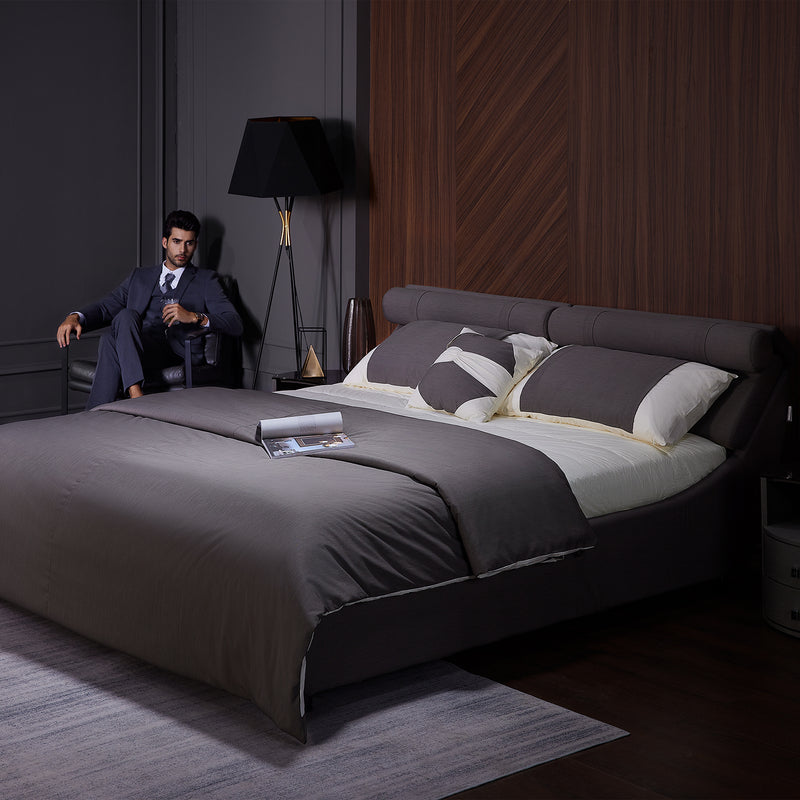 DeRUCCI Bed Frame BZZ4 - 093C with dark gray bedding and headboard, nightstand with lamp, and a man in a suit seated beside the bed in a modern bedroom setting.